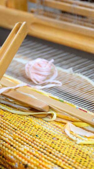 keltainen räsymatto kangaspuissa, kutojan kädet ja vaalea kude ja sukkula käsissä